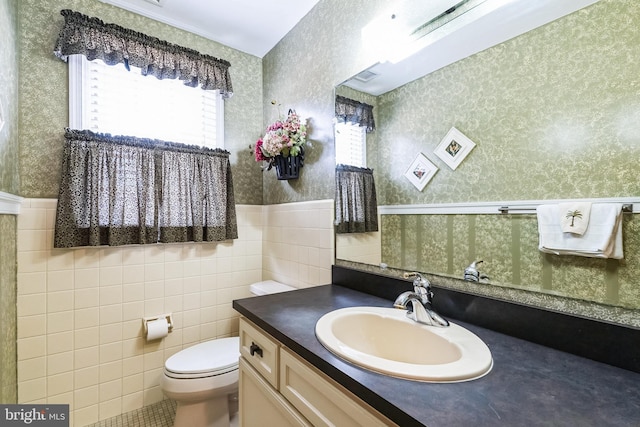 bathroom featuring tile walls, toilet, wainscoting, vanity, and wallpapered walls