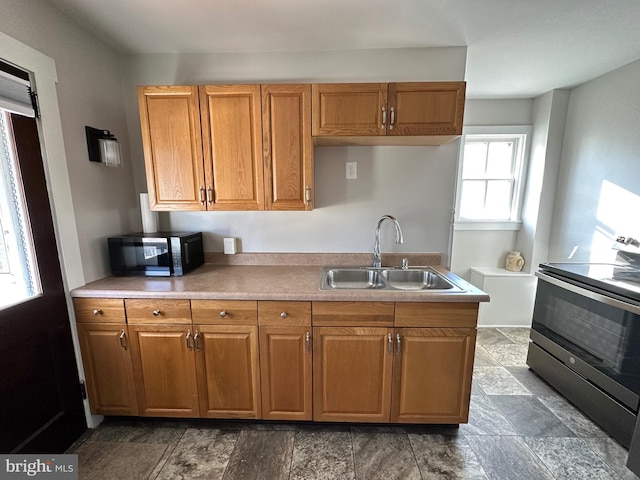 kitchen featuring light countertops, stainless steel range with electric stovetop, brown cabinetry, stone finish floor, and a sink