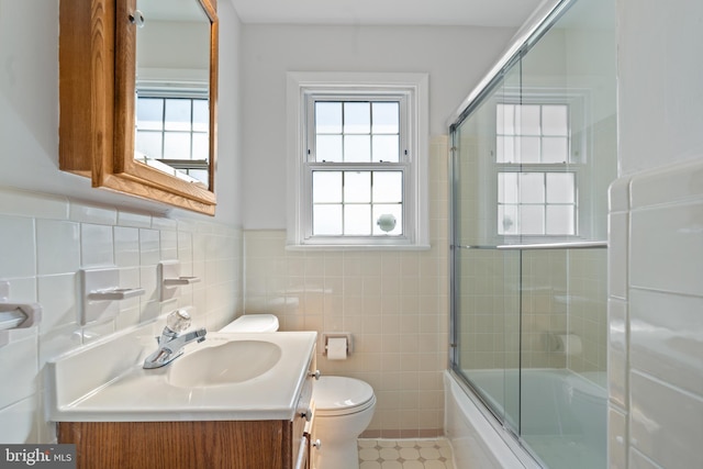 bathroom with toilet, shower / bath combination with glass door, vanity, tile walls, and wainscoting