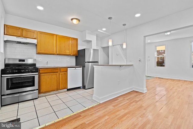 kitchen featuring hanging light fixtures, decorative backsplash, appliances with stainless steel finishes, brown cabinetry, and under cabinet range hood