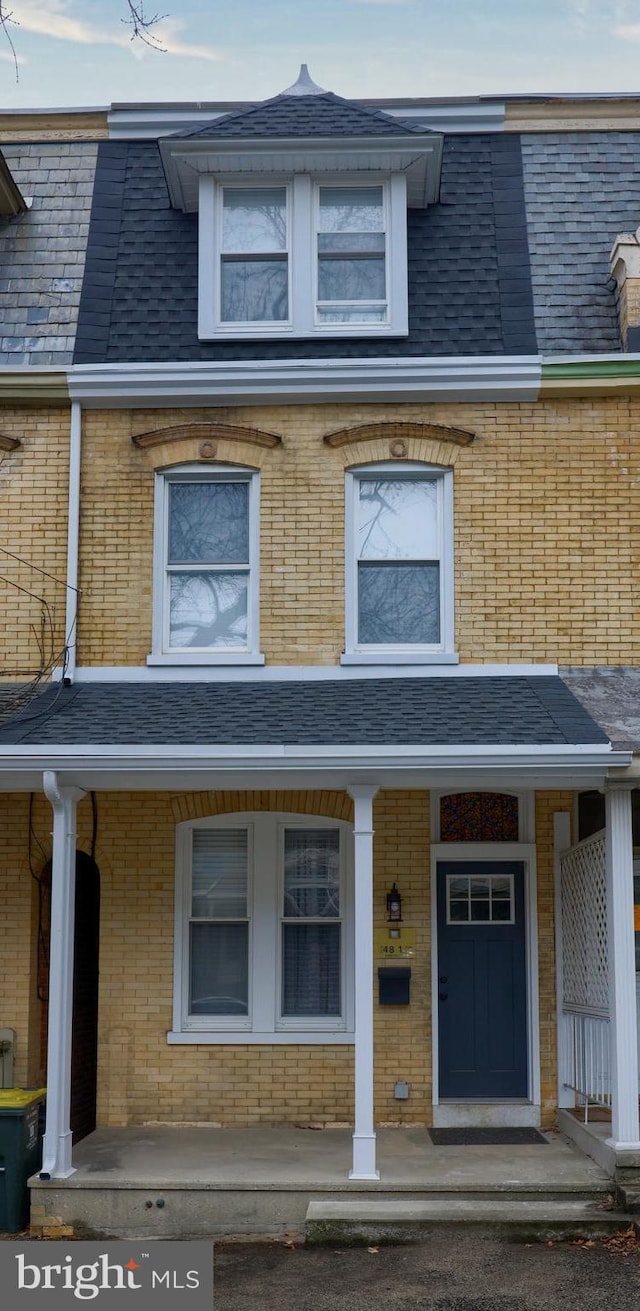 view of front facade featuring a shingled roof, covered porch, brick siding, and mansard roof