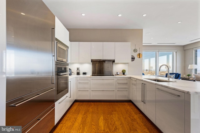 kitchen with light countertops, white cabinetry, a sink, built in appliances, and a peninsula