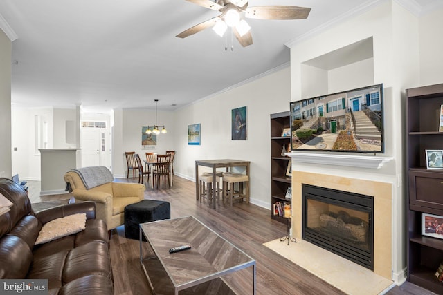 living room featuring ceiling fan with notable chandelier, wood finished floors, baseboards, ornamental molding, and a glass covered fireplace