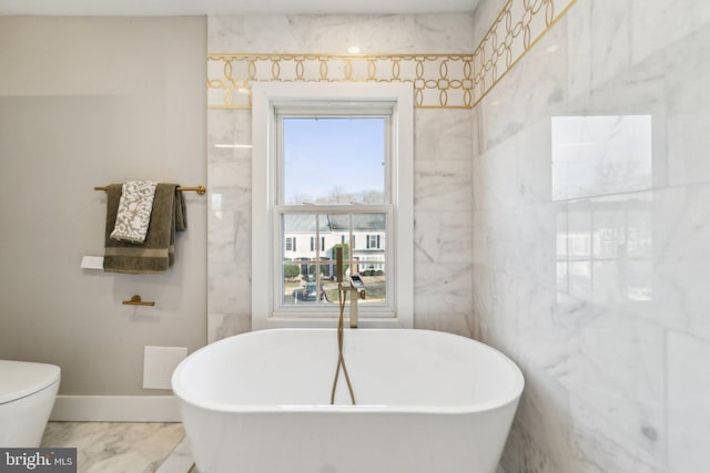 bathroom featuring marble finish floor, tile walls, toilet, a freestanding tub, and baseboards