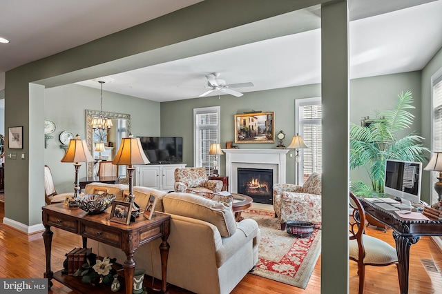 living area featuring ceiling fan with notable chandelier, light wood-type flooring, a glass covered fireplace, and baseboards