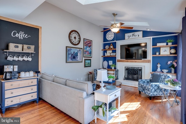 living room featuring lofted ceiling with skylight, a brick fireplace, a ceiling fan, and wood finished floors