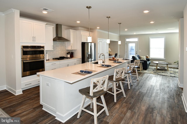 kitchen featuring dark wood finished floors, appliances with stainless steel finishes, white cabinets, wall chimney range hood, and a large island with sink