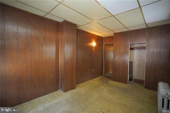 corridor with a paneled ceiling, carpet flooring, wooden walls, and radiator heating unit