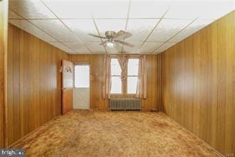 spare room with a paneled ceiling, wooden walls, a ceiling fan, radiator heating unit, and carpet