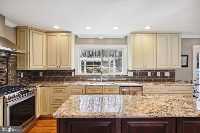 kitchen featuring light stone countertops, a sink, stainless steel appliances, cream cabinetry, and wall chimney range hood