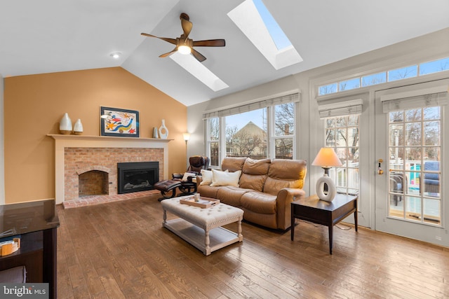 living room with ceiling fan, vaulted ceiling with skylight, a brick fireplace, and wood-type flooring