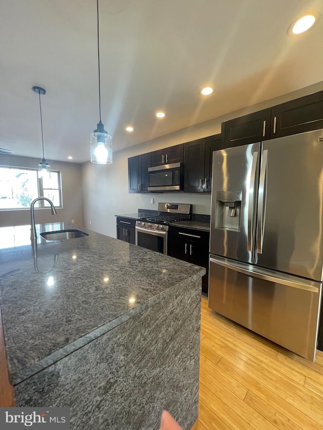 kitchen with light wood-type flooring, dark cabinets, a sink, dark stone countertops, and appliances with stainless steel finishes
