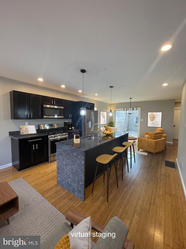 kitchen with a kitchen bar, appliances with stainless steel finishes, dark cabinetry, light wood-style flooring, and an island with sink