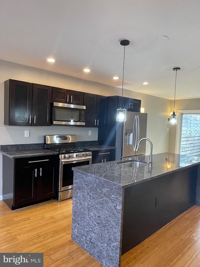 kitchen with hanging light fixtures, a sink, light wood finished floors, dark stone counters, and stainless steel appliances