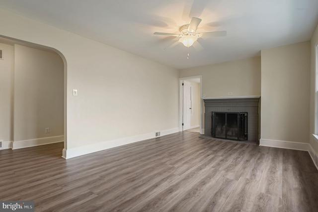 unfurnished living room featuring arched walkways, a brick fireplace, ceiling fan, and wood finished floors