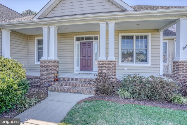 view of exterior entry with a shingled roof, a porch, and brick siding