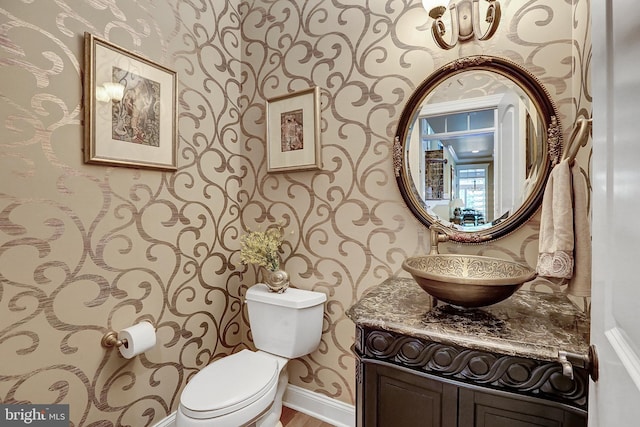 half bathroom featuring toilet, wallpapered walls, baseboards, and vanity