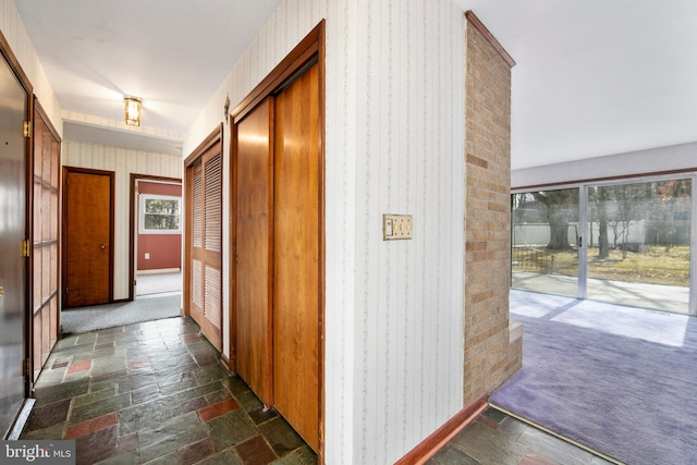 hall featuring dark colored carpet, stone tile flooring, baseboards, and wallpapered walls