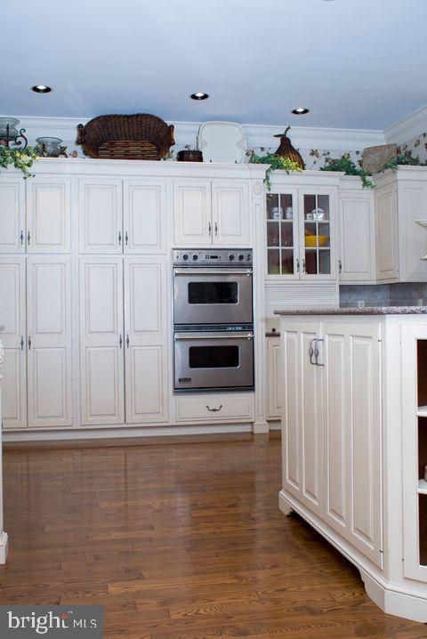 kitchen with dark wood finished floors, crown molding, glass insert cabinets, stainless steel double oven, and white cabinetry