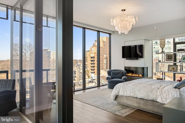 bedroom with expansive windows, a warm lit fireplace, wood finished floors, and a chandelier