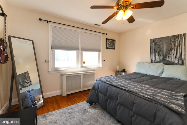 bedroom featuring ceiling fan, wood finished floors, visible vents, baseboards, and radiator heating unit