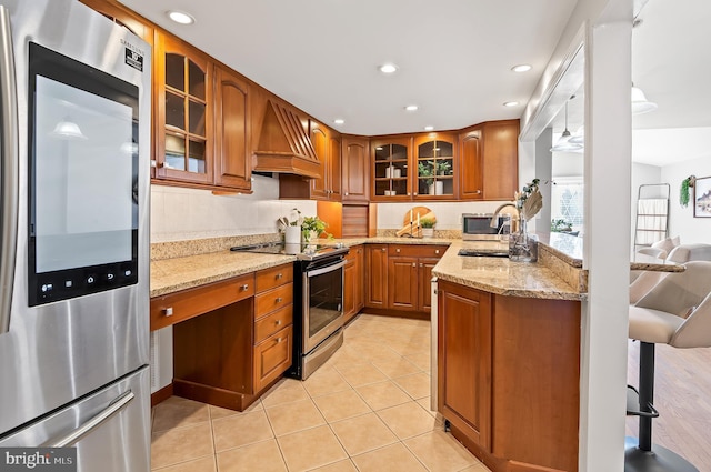 kitchen featuring custom range hood, appliances with stainless steel finishes, brown cabinetry, a sink, and light stone countertops