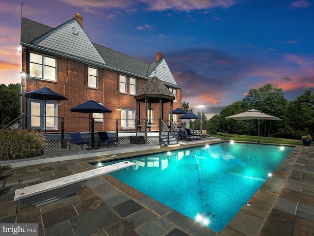 pool at dusk featuring an outdoor pool, a diving board, and a patio area