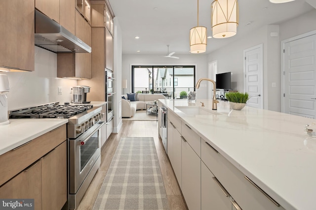 kitchen with light wood finished floors, high end stove, under cabinet range hood, open floor plan, and a sink