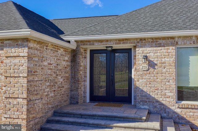 view of exterior entry with french doors, brick siding, and roof with shingles