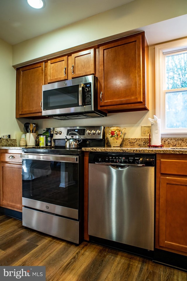 kitchen with brown cabinets, dark wood-style floors, light stone counters, and stainless steel appliances