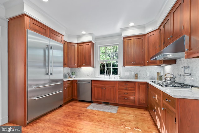 kitchen featuring light wood-style flooring, appliances with stainless steel finishes, brown cabinetry, a sink, and under cabinet range hood