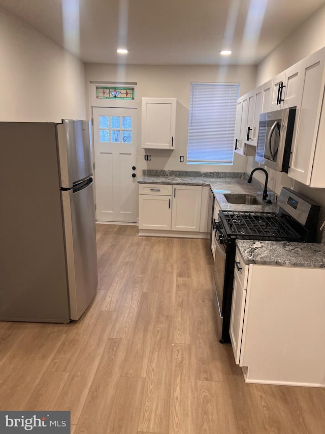 kitchen featuring stainless steel appliances, a sink, white cabinets, light wood-type flooring, and dark stone counters