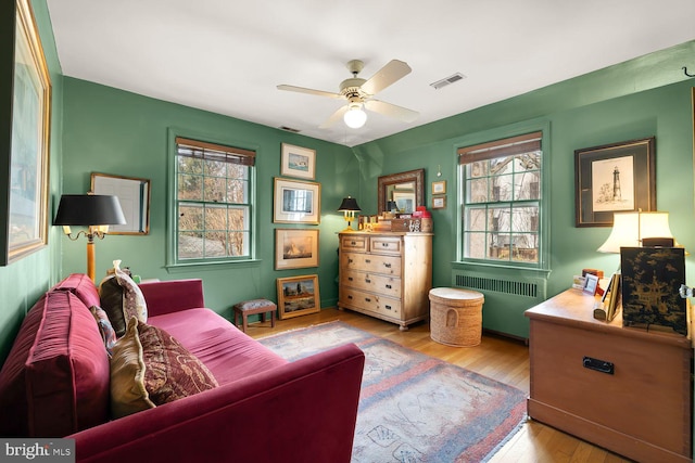 living area with a ceiling fan, light wood-type flooring, visible vents, and radiator heating unit