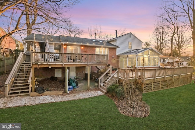 back of house at dusk featuring a shingled roof, a wooden deck, stairs, a yard, and a sunroom