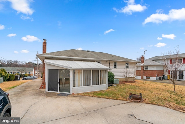 back of property with a sunroom, a yard, concrete driveway, a shingled roof, and a chimney