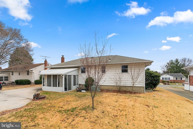 rear view of house with a shingled roof, a lawn, a chimney, a sunroom, and driveway