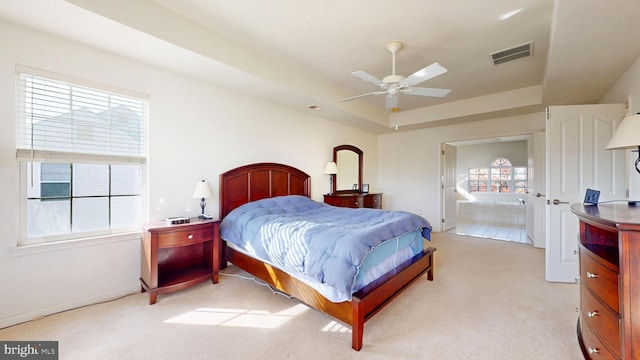 bedroom with a tray ceiling, light colored carpet, visible vents, connected bathroom, and ceiling fan