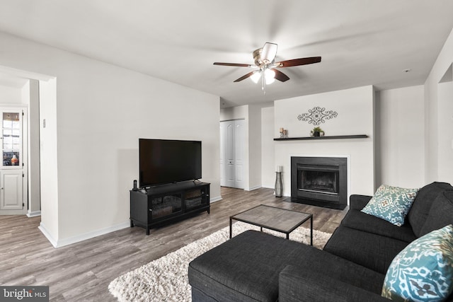 living room featuring a fireplace with flush hearth, wood finished floors, baseboards, and ceiling fan