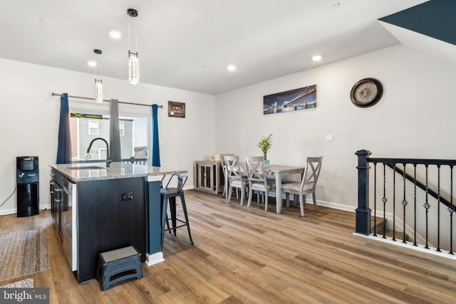 kitchen with a kitchen island with sink, wood finished floors, a sink, light stone countertops, and pendant lighting