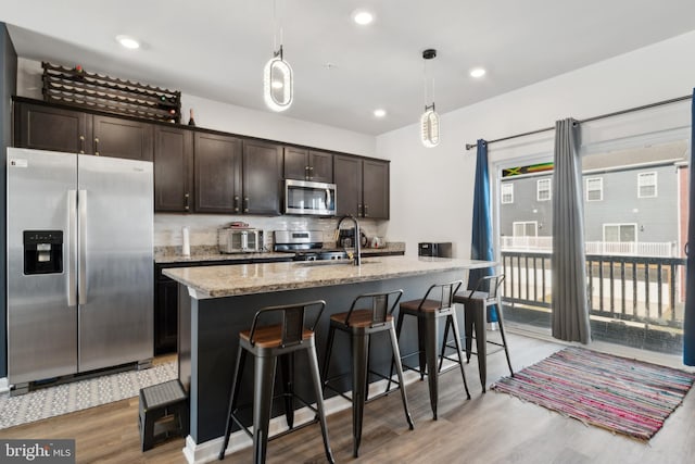 kitchen featuring appliances with stainless steel finishes, light stone counters, hanging light fixtures, a kitchen island with sink, and dark brown cabinets