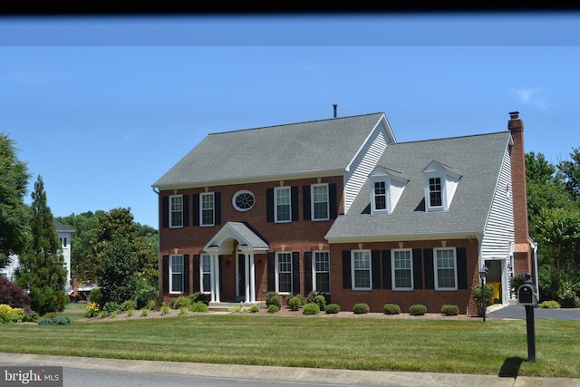 view of front of house with a shingled roof, a front yard, brick siding, and a chimney