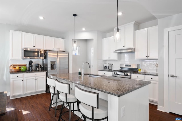 kitchen with under cabinet range hood, a sink, white cabinets, appliances with stainless steel finishes, and a center island with sink