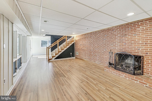 unfurnished living room with a paneled ceiling, a brick fireplace, stairs, and wood finished floors