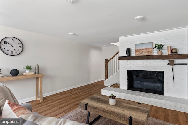 living area featuring a brick fireplace, stairway, baseboards, and wood finished floors