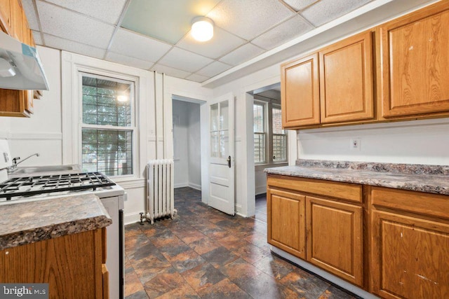 kitchen featuring white gas stove, a drop ceiling, under cabinet range hood, radiator, and dark countertops