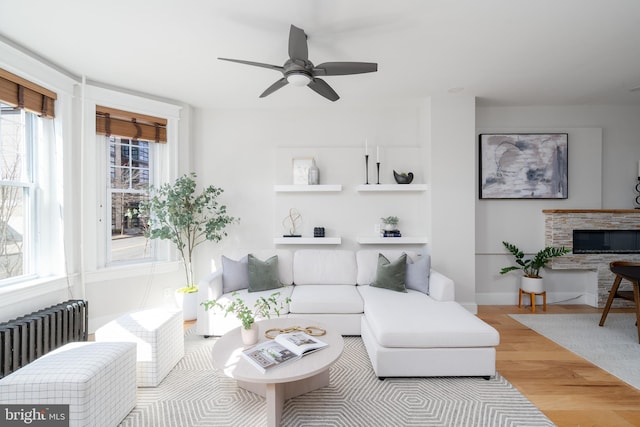 living room featuring built in features, radiator heating unit, a ceiling fan, a stone fireplace, and wood finished floors