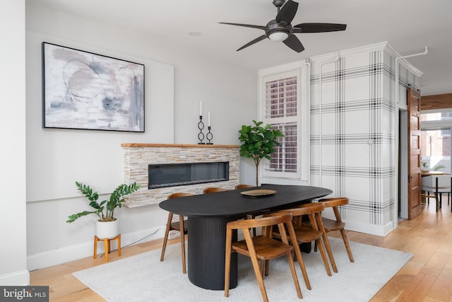 dining area featuring light wood-style flooring, ceiling fan, a stone fireplace, and baseboards