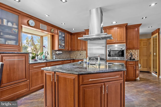 kitchen featuring stainless steel oven, brown cabinetry, a kitchen island with sink, and island exhaust hood