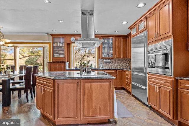 kitchen featuring a kitchen island, stainless steel built in fridge, dark stone counters, island exhaust hood, and glass insert cabinets