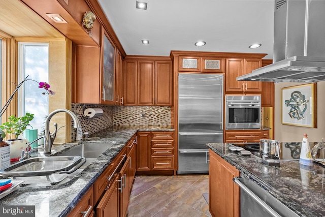 kitchen featuring island range hood, glass insert cabinets, appliances with stainless steel finishes, brown cabinets, and a sink